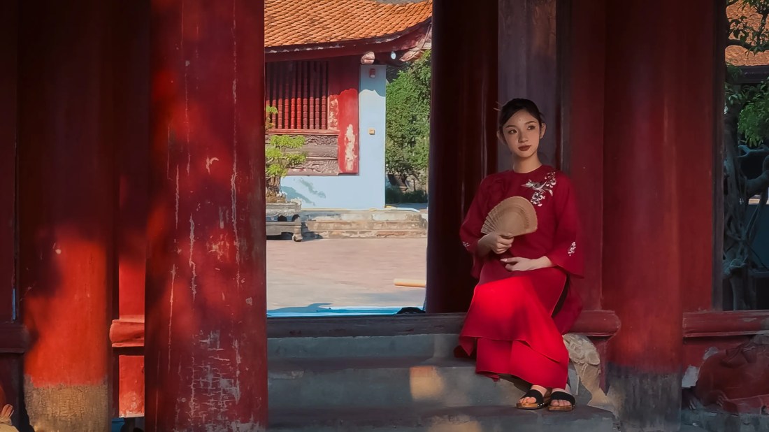Young lady in traditional Vietnamese dress sitting with a fan by a courtyard