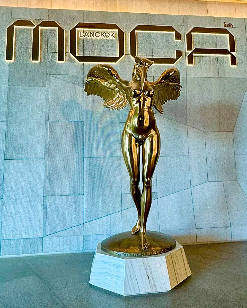 a gold human figure with eagle head and wings statue in front of the MOCA Bangkok sign at the museum